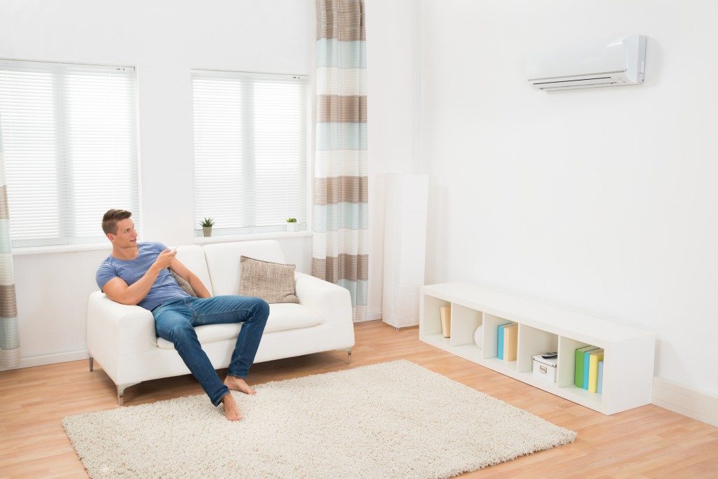 Man Sitting On Sofa Operating Air Conditioner With Remote Controller